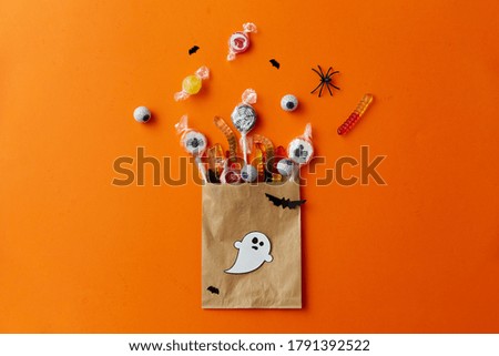 halloween, sweets and holiday concept - candies in paper bag and decorations on orange background