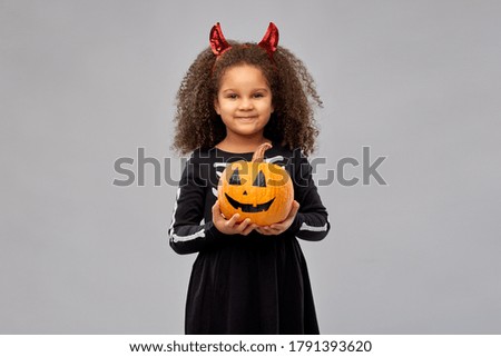 halloween, holiday and childhood concept - smiling african american girl in black costume dress with skeleton bones and red devil's horns holding jack-o-lantern pumpkin over grey background