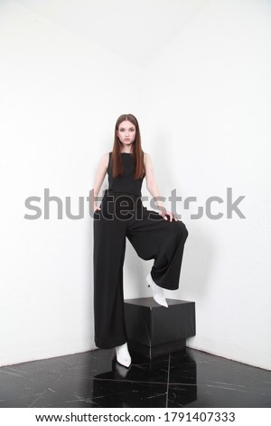 Serious young woman  in a black blouse, wide pants and white leather boots. Beauty, trendy, ladies fashion. Full length studio shot isolated on white wall background with black floor. Copy space