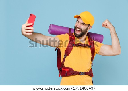 Cheerful traveler young man with backpack isolated on blue background. Tourist traveling on weekend getaway. Tourism discovering hiking concept. Doing selfie shot on mobile phone doing winner gesture