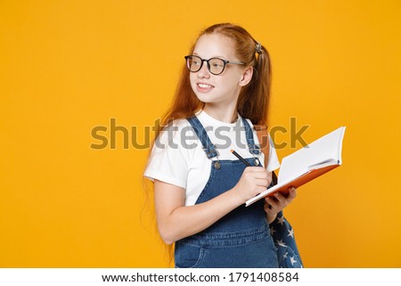Young redhead girl 12-13 years old in white t-shirt denim uniform eyeglasses backpack hold school notebook book isolated on yellow background children studio portrait Kids education lifestyle concept.