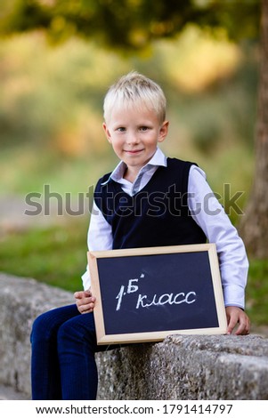 
A boy with blond hair in a school uniform sits in an autumn park with a sign "Grade 1 B"
