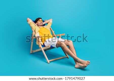 Full length profile side photo of positive guy traveler relax rest beach sun bathing lie comfort deckchair stretch hands wear white shorts isolated over blue color background Royalty-Free Stock Photo #1791479027