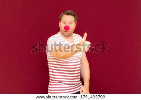 The gesturing charming trendy and smiling man shows thumb up on red or burgundy wall.Red nose day or april fools day. The clown, fun, party, celebration, funny, joy, holiday, humor concept