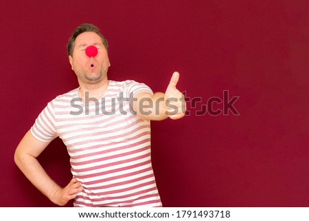 The self-satisfied trendy and smiling man shows thumb up on red or burgundy wall.Red nose day or april fools day. The clown, fun, party, celebration, funny, joy, holiday, humor concept