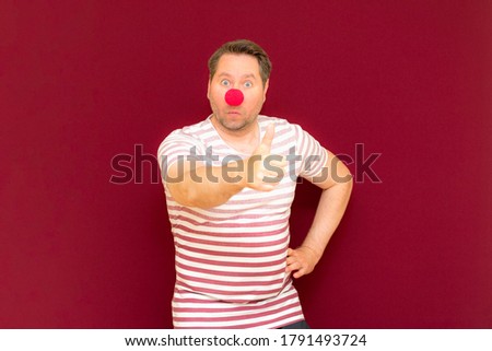 The gesturing charming trendy and smiling man shows thumb up on red or burgundy wall.Red nose day or april fools day. The clown, fun, party, celebration, funny, joy, holiday, humor concept