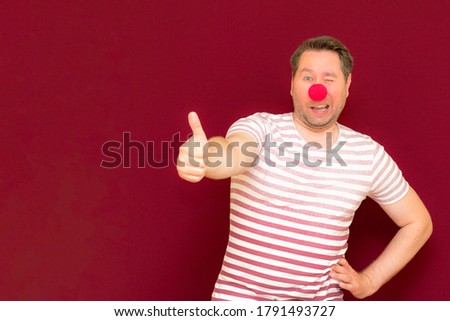 The self-satisfied trendy and smiling man shows thumb up on red or burgundy wall.Red nose day or april fools day. The clown, fun, party, celebration, funny, joy, holiday, humor concept