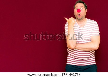 The happy surprised charming trendy and smiling manshows to the side or copy space burgundy wall.Red nose day or april fools day. The clown, fun, party, celebration, funny, joy, holiday,humor concept