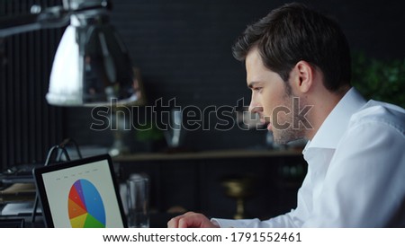 Focused businessman looking at graphs on laptop computer in office interior. Male freelancer analyzing financial reports on laptop screen. Serious business man working on laptop at home office