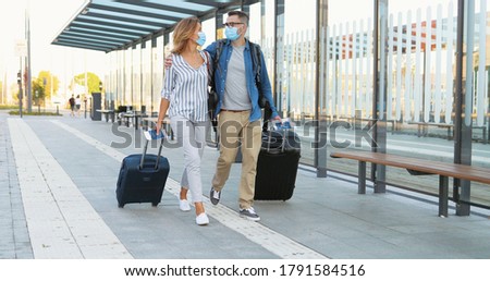 Caucasian happy young married couple in love walking at bus stop and carrying suitcases on wheels. Beautiful woman and handsome man in medical masks hugging and travelling during covid-19 pandemic.