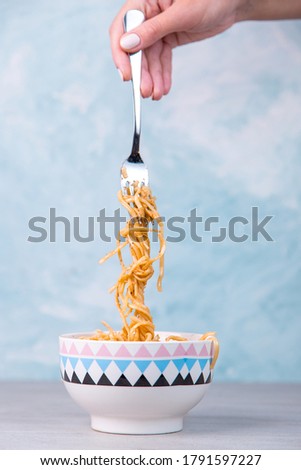 Noodles with sauce in colored bowl, hand holds a fork of pasta hanging, appetite spaghetti on a blue background.