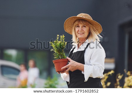 Holding pot in hands. Senior woman is in the mini garden at daytime. Building exterior behind.
