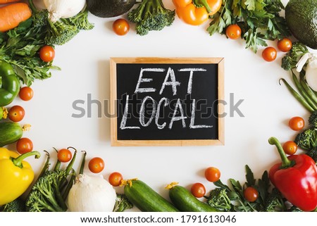 Small blackboard with handwritten text "Eat Local" and assorted seasonal vegetables on white background, top view. Eat local food concept