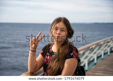 Photo of a young preteen girl with long brown hair makes a v-sign towards the camera. Blue ocean and sky background