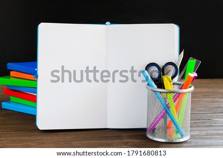 Back to school. Open empty notebook, stack of books and school stationery on black board background.