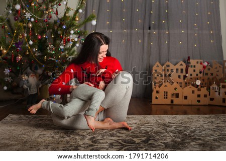 Mom and daughter in a red sweater for Christmas at the Christmas tree. A girl and her mother are hugging, kissing at the Christmas tree at home for the New Year 