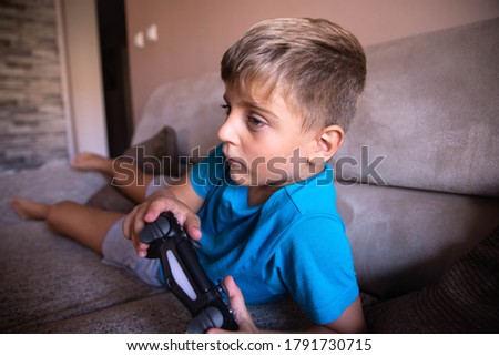 A beautiful little boy is lying on the couch and playing video games