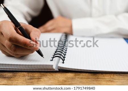 A schoolgirl, dressed in a white blouse, sits at a wooden desk and holds in her hand a black pen over a notebook. School concept.