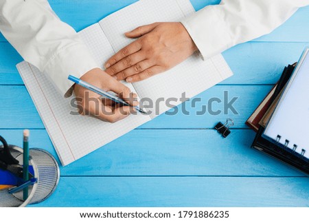 A schoolgirl, dressed in a white dress shirt, sits at a blue wooden table with school supplies and holds a pen over a notebook. View from above.