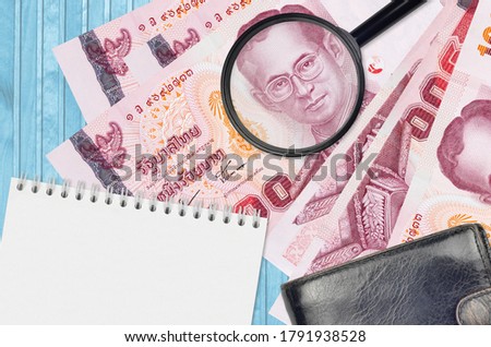 100 Thai Baht bills and magnifying glass with black purse and notepad. Concept of counterfeit money. Search for differences in details on money bills to detect fake
