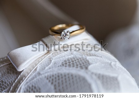 Close up picture of the new golden beautiful diamond engagement ring with clear round diamond placed on the bow of cream lace vintage wedding shoes, dark background