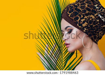 Summer sea and the hot weather, the beach trend. A young beautiful multi-ethnic woman in a leopard turban and yellow bathing suit looks out from behind a palm branch, yellow mango background