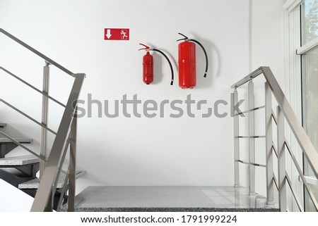 Fire extinguishers and emergency exit sign on white wall near staircase indoors