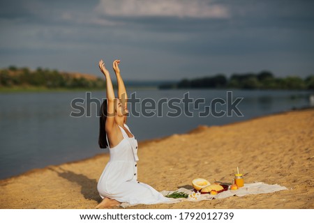Young woman in white dress sitting on the sandy beach with hands up and enjoying summer picnic.  Food on the shore. She is resting on the beach.