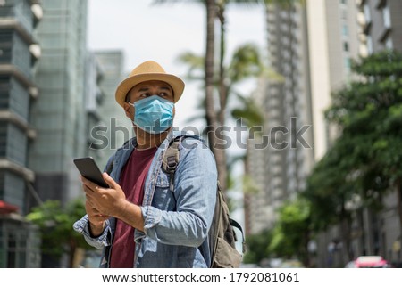 Young asian backpacker traveling in urban city using smartphone to search the location to travel. He wearing face mask for protect virus safety travel.