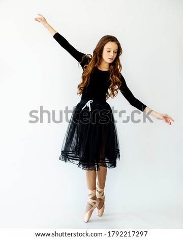 A young beautiful ballerina girl stands in motion on Pointe shoes in a black swimsuit and skirt with light background. Full-length photo. Studio of modern choreography, ballet, dance