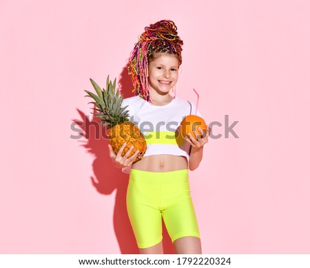 cute little girl with multi-colored pigtails on her head smiles happily, holding a pineapple and an orange in her hands. Cropped portrait isolated on pink, copy space. Childhood, emotions, summer