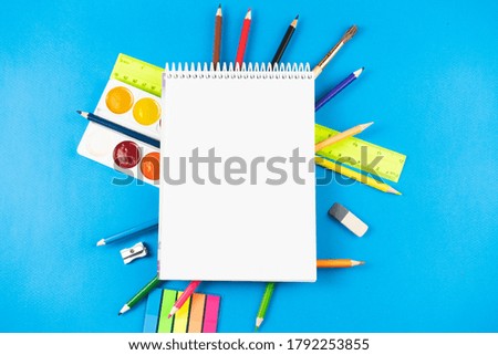 On the colorful school supplies lay a white blank notebook on a blue pastel background. Place for text.