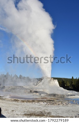 A picture of an active geyser in Yellow Stone National Park with a rainbow in the sky. The rainbow is seen from the water being expelled from the geyser. There is steam and water seen in the blue sky.