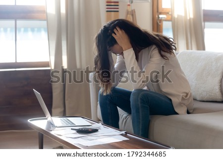 Stressed young woman holding head in hands, depressed about financial problems, sitting on sofa in living room. Unhappy millennial girl feeling hopeless managing monthly budget, bankruptcy concept. Royalty-Free Stock Photo #1792344685