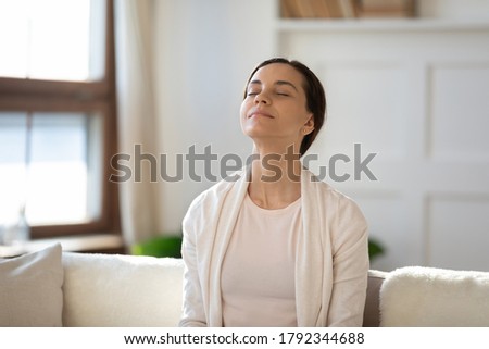Mindful young woman breathing fresh air with closed eyes, sitting on sofa alone indoors. Peaceful happy millennial girl meditating in living room, enjoying affirmations or lazy free weekend time. Royalty-Free Stock Photo #1792344688