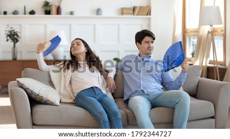 Stressed tired young family couple sitting together on sofa, fanning fresh cool air with paper appliances, feeling unwell at summertime, suffering from sunstroke or high temperature at home. Royalty-Free Stock Photo #1792344727