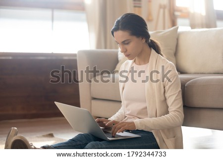 Focused young female freelancer sitting on floor leaning on sofa, working remotely on computer in living room. Serious woman web surfing information or studying online from home, using laptop.