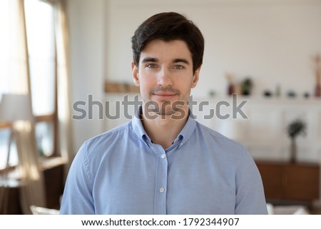Portrait of smiling young handsome man freelancer standing in modern living room. Happy millennial bearded guy homeowner looking at camera, posing at home, social network profile picture concept.