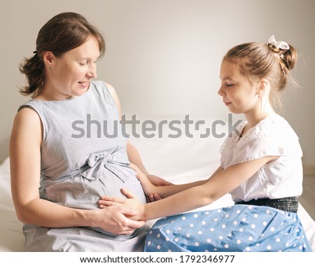 Beautiful pregnant woman and her cute little daughter are smiling while spending time together at home.