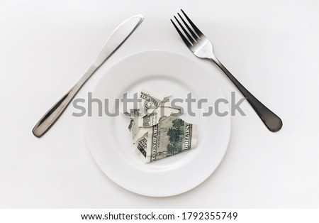 A hundred-dollar bill lies on a ceramic plate between a fork and a knife on a white tablecloth. The concept of business lunch, banking, life rentier and financial well-being.