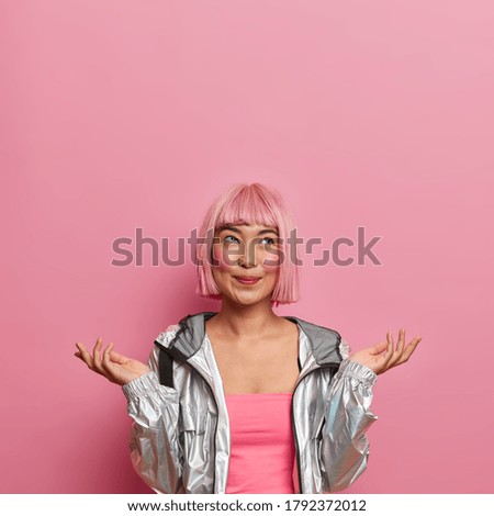 Ethnic woman with pink hairstyle, raises palms and shrugs shoulders, smiles positively and looks above, cannot decide, dressed in glittering silver jacket, isolated on pink background with copy space
