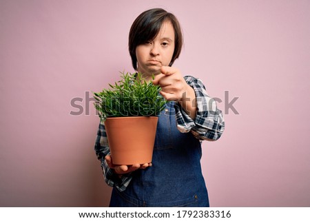 Young down syndrome gardener woman wearing worker apron holding green plant pot pointing with finger to the camera and to you, hand sign, positive and confident gesture from the front