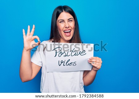 Young beautiful woman asking for optimist attitude holding paper with positive vibes message doing ok sign with fingers, smiling friendly gesturing excellent symbol
