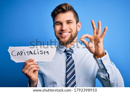 Young blond businessman with beard and blue eyes holding paper with capitalism message doing ok sign with fingers, excellent symbol