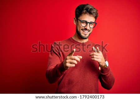 Young handsome man with beard wearing glasses and sweater standing over red background pointing fingers to camera with happy and funny face. Good energy and vibes.