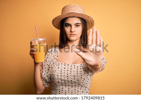 Young beautiful brunette woman on vacation wearing summer hat drinking orange juice with open hand doing stop sign with serious and confident expression, defense gesture