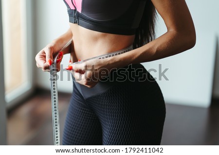 Fitness motivation and successful weight loss. Woman with perfect slim body measuring her waistline with tape, close up. Diet, home training, healthy lifestyle concept Royalty-Free Stock Photo #1792411042