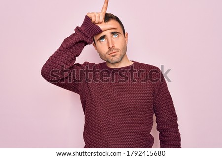 Young handsome man with blue eyes wearing casual sweater standing over pink background making fun of people with fingers on forehead doing loser gesture mocking and insulting.