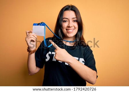Young brunette worker woman wearing staff t-shirt as uniform showing id card very happy pointing with hand and finger