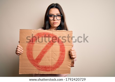 Young brunette woman holding protest banner with prohibited symbol over isolated background with a confident expression on smart face thinking serious